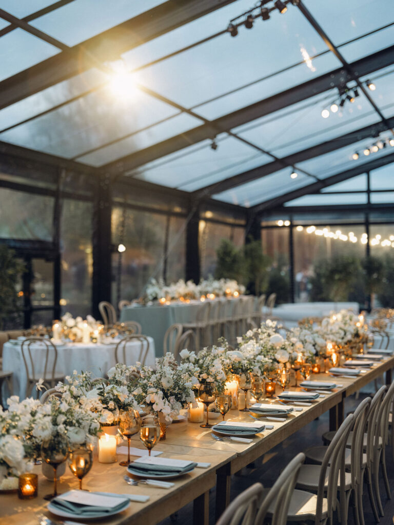 A moody, garden-inspired wedding reception featuring elegant white and green floral centerpieces under a glass tent for fall wedding. Cloud roses, ranunculus, lisianthus, chamomile, queen anne’s lace, saponaria, delphinium, flannel flower and delicate greenery create a heavy floral focal point for tables in tones of white, cream and green with amber table accents and warm candle lighting. Lush greenery drapes across the ceiling. Design by Rosemary and Finch Floral Design in Nashville, TN. 