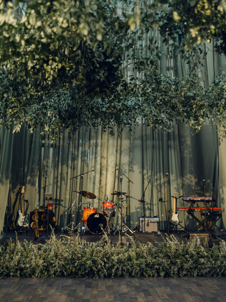 Lush suspended greenery creates a dramatic garden canopy above candlelit tables filled with elegant white and green floral centerpieces. The hanging foliage installation brings organic movement and depth, casting whimsical shadows throughout the tent and dance floor. The installation is surrounded by gorgeous centerpieces of cream, white and green tones with amber table accents scattered around the room. Design by Rosemary and Finch Floral Design in Nashville, TN.