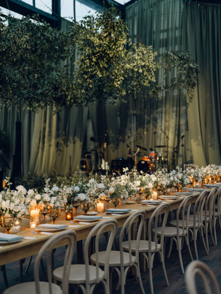 A moody, garden-inspired wedding reception featuring elegant white and green floral centerpieces under a glass tent for fall wedding. Cloud roses, ranunculus, lisianthus, chamomile, queen anne’s lace, saponaria, delphinium, flannel flower and delicate greenery create a heavy floral focal point for tables in tones of white, cream and green with amber table accents and warm candle lighting. Lush greenery drapes across the ceiling. Design by Rosemary and Finch Floral Design in Nashville, TN. 