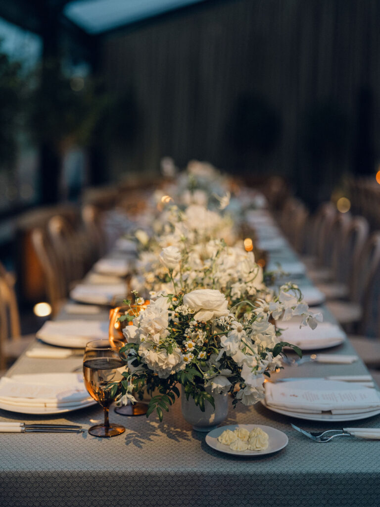 A moody, garden-inspired wedding reception featuring elegant white and green floral centerpieces under a glass tent for fall wedding. Cloud roses, ranunculus, lisianthus, chamomile, queen anne’s lace, saponaria, delphinium, flannel flower and delicate greenery create a heavy floral focal point for tables in tones of white, cream and green with amber table accents and warm candle lighting. Lush greenery drapes across the ceiling. Design by Rosemary and Finch Floral Design in Nashville, TN. 