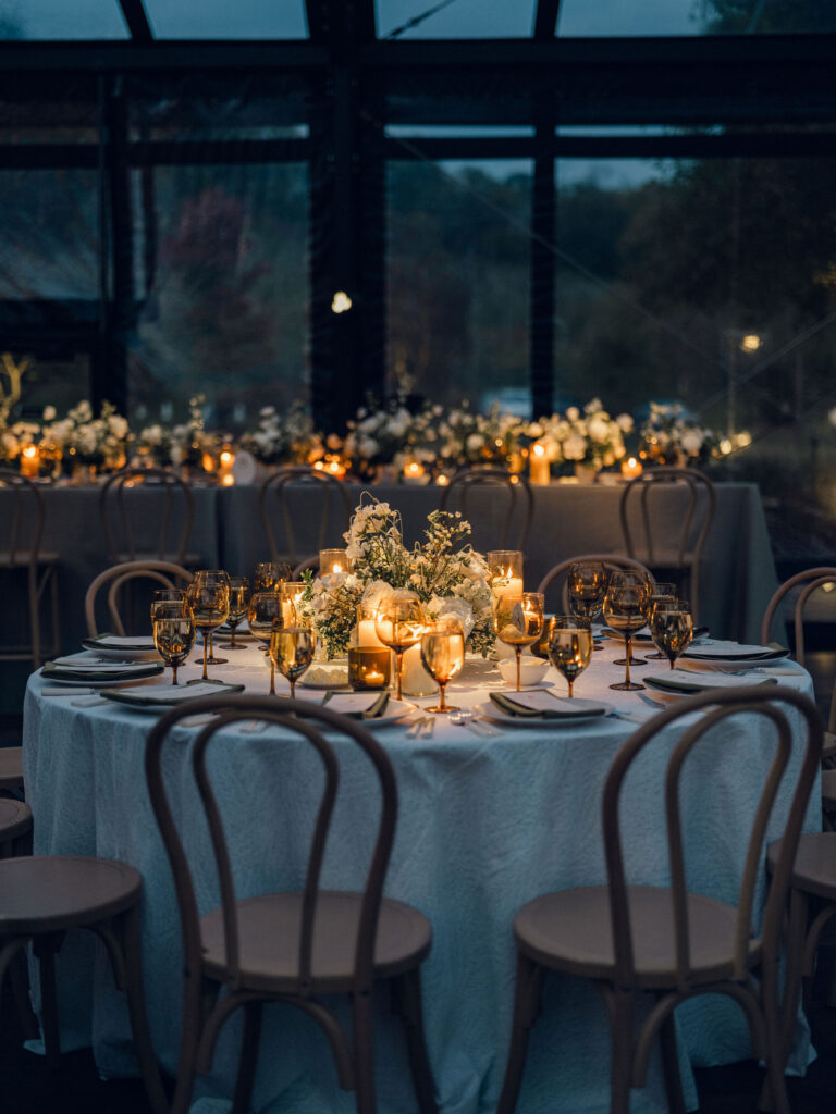 A moody, garden-inspired wedding reception featuring elegant white and green floral centerpieces under a glass tent for fall wedding. Cloud roses, ranunculus, lisianthus, chamomile, queen anne’s lace, saponaria, delphinium, flannel flower and delicate greenery create a heavy floral focal point for tables in tones of white, cream and green with amber table accents and warm candle lighting. Lush greenery drapes across the ceiling. Design by Rosemary and Finch Floral Design in Nashville, TN. 