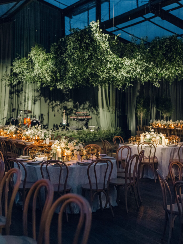 Lush suspended greenery creates a dramatic garden canopy above candlelit tables filled with elegant white and green floral centerpieces. The hanging foliage installation brings organic movement and depth, casting whimsical shadows throughout the tent and dance floor. The installation is surrounded by gorgeous centerpieces of cream, white and green tones with amber table accents scattered around the room. Design by Rosemary and Finch Floral Design in Nashville, TN.