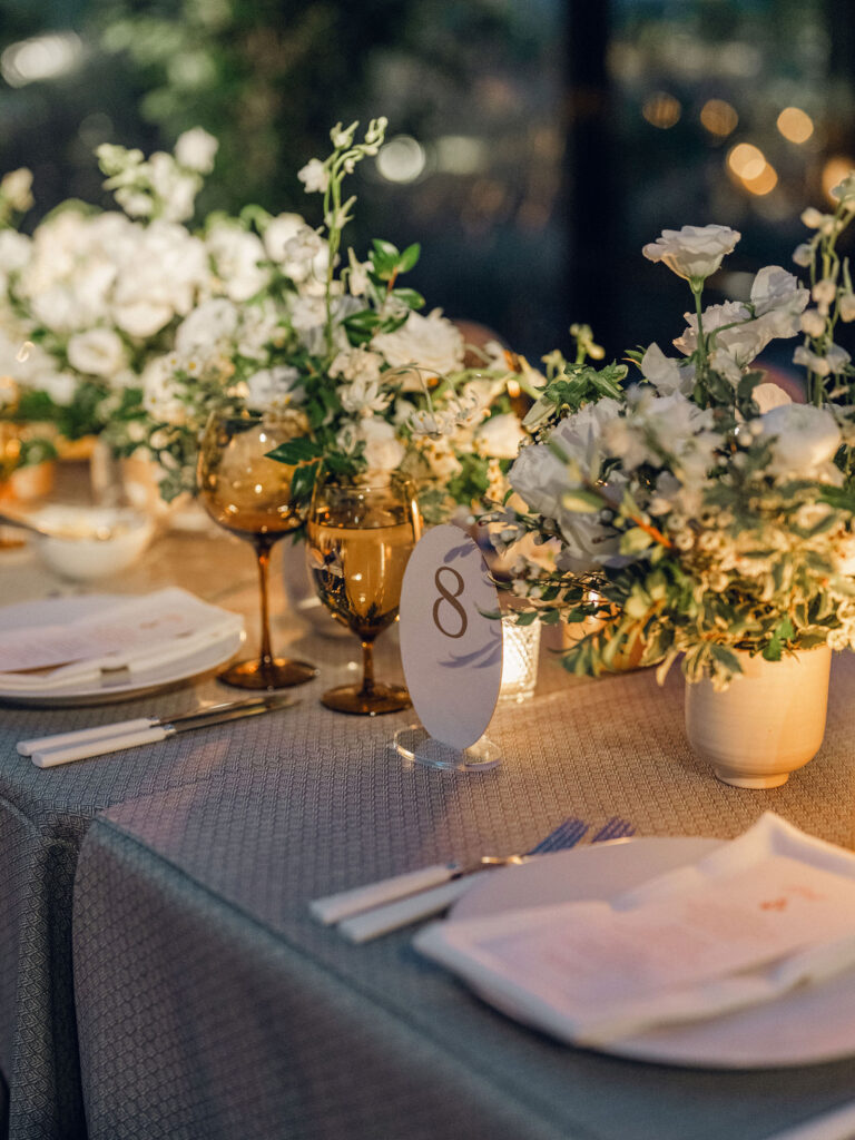A moody, garden-inspired wedding reception featuring elegant white and green floral centerpieces under a glass tent for fall wedding. Cloud roses, ranunculus, lisianthus, chamomile, queen anne’s lace, saponaria, delphinium, flannel flower and delicate greenery create a heavy floral focal point for tables in tones of white, cream and green with amber table accents and warm candle lighting. Lush greenery drapes across the ceiling. Design by Rosemary and Finch Floral Design in Nashville, TN. 