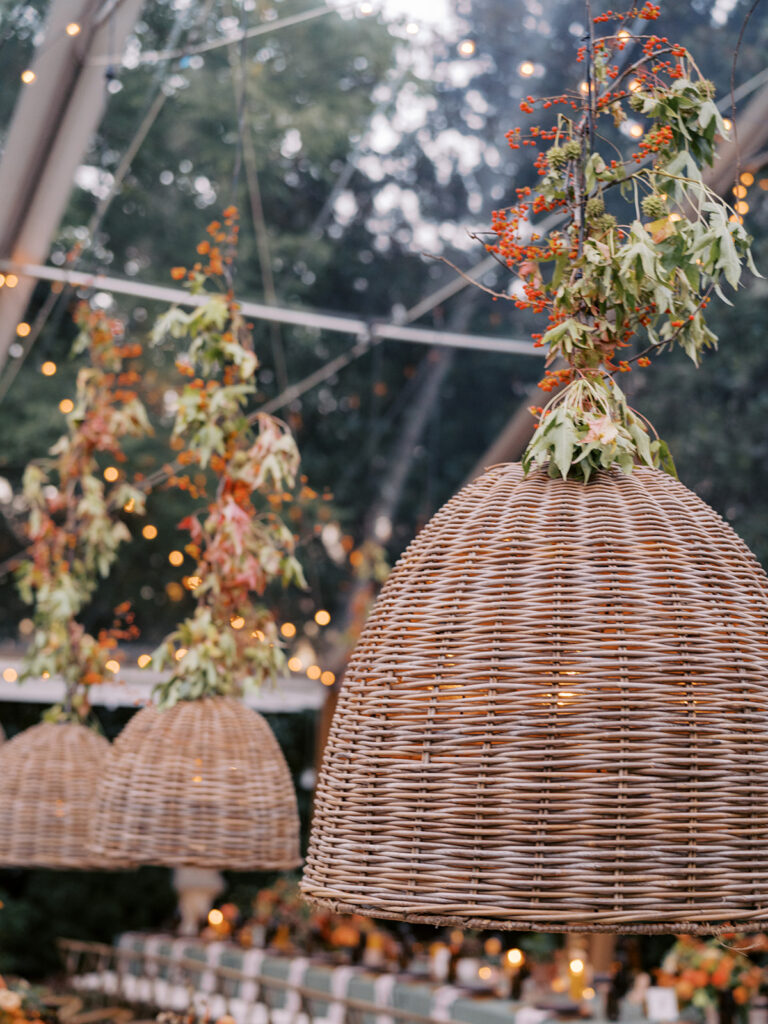 Elegant, rustic pendants with branchy greenery hang above long tables for a private birthday party in Nashville, TN. Fall foliage combined with bittersweet are wrapped around each pendant to create an autumnal and warm atmosphere. The warm lighting and greenery complement stunning table arrangements and the warm glow from votives scattered throughout the reception. Design by Rosemary and Finch Floral Design in Nashville, TN. 