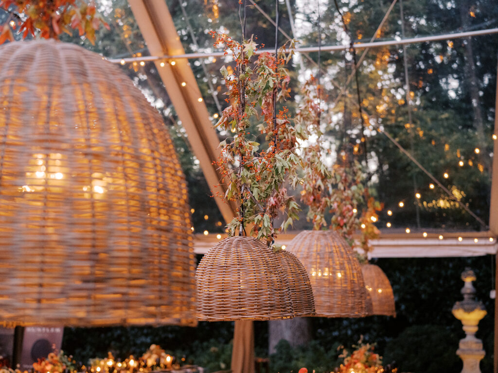 Elegant, rustic pendants with branchy greenery hang above long tables for a private birthday party in Nashville, TN. Fall foliage combined with bittersweet are wrapped around each pendant to create an autumnal and warm atmosphere. The warm lighting and greenery complement stunning table arrangements and the warm glow from votives scattered throughout the reception. Design by Rosemary and Finch Floral Design in Nashville, TN. 