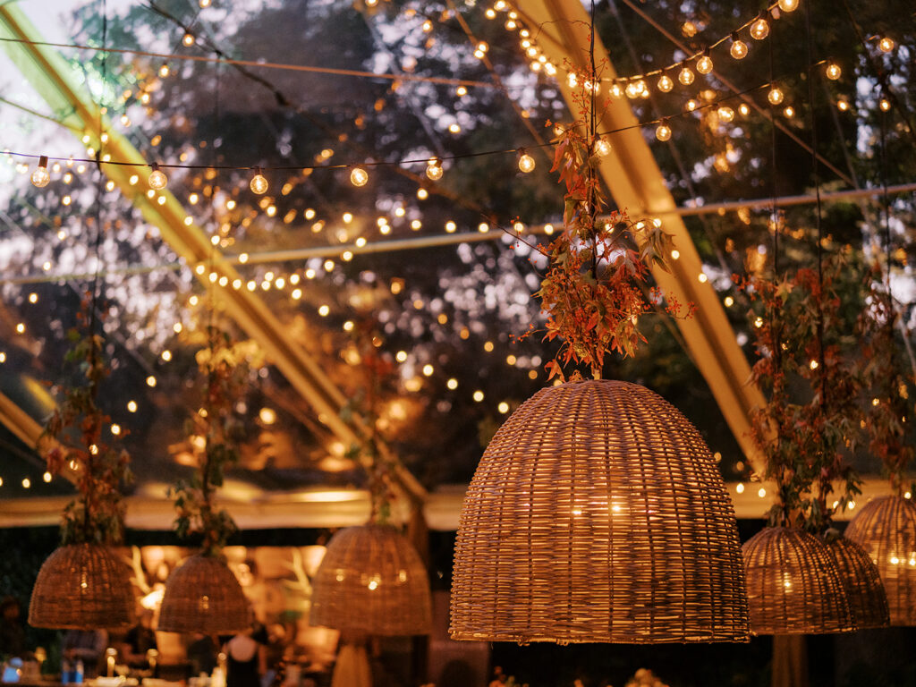 Elegant, rustic pendants with branchy greenery hang above long tables for a private birthday party in Nashville, TN. Fall foliage combined with bittersweet are wrapped around each pendant to create an autumnal and warm atmosphere. The warm lighting and greenery complement stunning table arrangements and the warm glow from votives scattered throughout the reception. Design by Rosemary and Finch Floral Design in Nashville, TN. 