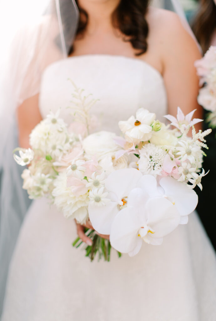Stunning, elegant flower-forward all-white bridal bouquet with hints of blush and pink for Belle Meade wedding in Nashville. Florals highlighted are phalaenopsis, dahlias, garden roses, majolika roses, ranunculus, butterfly ranunculus, clematis, sweet peas, astilbe, scabiosa and light greenery. The white phalaenopsis creates a soft and bright focal moment for the bouquet. Design by Rosemary and Finch Floral Design in Nashville, TN. 
