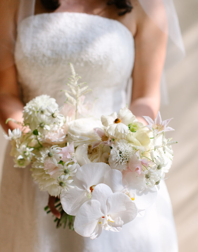 Stunning, elegant flower-forward all-white bridal bouquet with hints of blush and pink for Belle Meade wedding in Nashville. Florals highlighted are phalaenopsis, dahlias, garden roses, majolika roses, ranunculus, butterfly ranunculus, clematis, sweet peas, astilbe, scabiosa and light greenery. The white phalaenopsis creates a soft and bright focal moment for the bouquet. Design by Rosemary and Finch Floral Design in Nashville, TN. 