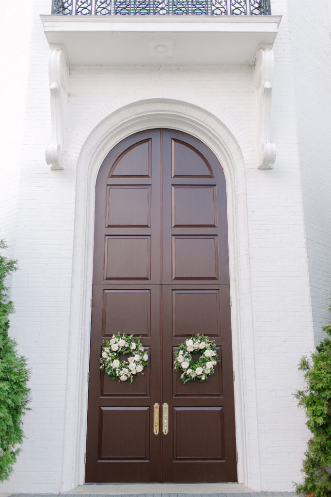 Wedding floral wreaths add a romantic, garden-inspired touch to private home and church doors for a wedding ceremony in Nashville. Designed with white florals and simple greenery, these handcrafted wreaths feature playa blanca roses, quicksand roses, champagne roses, majolikas,  and pieris, creating an elegant and timeless wedding floral statement. Design by Rosemary and Finch Floral Design in Nashville, TN. 