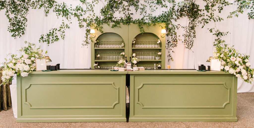 Stunning greenery is draped above and around the bar and reception area for a Nashville wedding at Hillwood Country Club. A wall of greenery adds to the elegance of the event and is accompanied by lush statement arrangements placed on the edge of the bar with all-white and blush florals consisting of dahlias, playa blancas, majolikas, garden roses, lisianthus, butterfly ranunculus, delphinium and more. Design by Rosemary and Finch Floral Design in Nashville, TN.