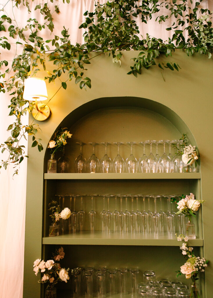 Stunning greenery is draped above and around the bar and reception area for a Nashville wedding at Hillwood Country Club. A wall of greenery adds to the elegance of the event and is accompanied by lush statement arrangements placed on the edge of the bar with all-white and blush florals consisting of dahlias, playa blancas, majolikas, garden roses, lisianthus, butterfly ranunculus, delphinium and more. Design by Rosemary and Finch Floral Design in Nashville, TN.