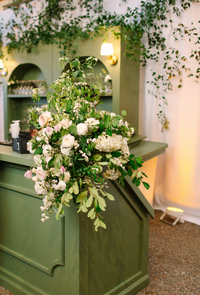 Stunning greenery is draped above the bar and reception area for a Nashville wedding at Hillwood Country Club. A wall of greenery adds to the elegance of the event and is accompanied by lush statement arrangements placed on the edge of the bar with all-white and blush florals consisting of dahlias, playa blancas, majolikas, garden roses, lisianthus, butterfly ranunculus, delphinium and more. Design by Rosemary and Finch Floral Design in Nashville, TN