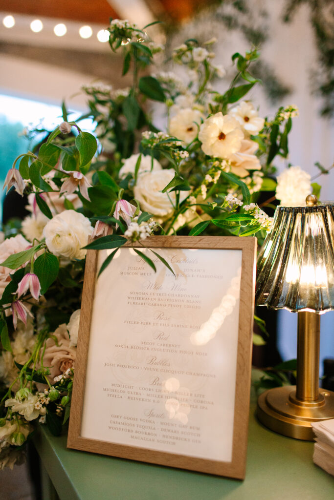 Stunning greenery is draped above the bar and reception area for a Nashville wedding at Hillwood Country Club. A wall of greenery adds to the elegance of the event and is accompanied by lush statement arrangements placed on the edge of the bar with all-white and blush florals consisting of dahlias, playa blancas, majolikas, garden roses, lisianthus, butterfly ranunculus, delphinium and more. Design by Rosemary and Finch Floral Design in Nashville, TN