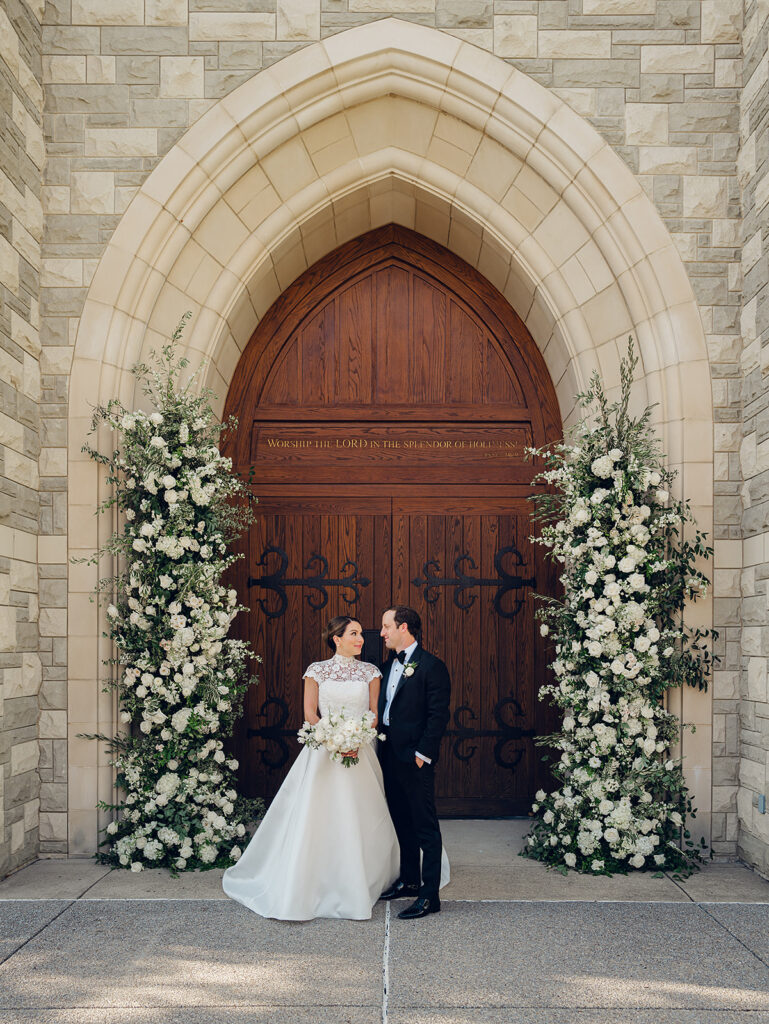 Stunning all-white floral arch for wedding ceremony at Covenant Presbyterian Church in Nashville. The arch creates an elegant focal point with flowers consisting of hydrangeas, cloud roses, lisianthus, ranunculus, tulips, dahlias, riceflower, stock, allium, delphinium, and ivy. Meadows with the same flowers surround the bottom of the arch on either side. Design by Rosemary & Finch Floral Design in Nashville, TN.