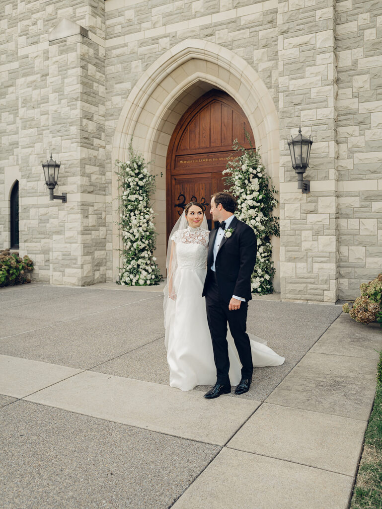 Stunning all-white floral arch for wedding ceremony at Covenant Presbyterian Church in Nashville. The arch creates an elegant focal point with flowers consisting of hydrangeas, cloud roses, lisianthus, ranunculus, tulips, dahlias, riceflower, stock, allium, delphinium, and ivy. Meadows with the same flowers surround the bottom of the arch on either side. Design by Rosemary & Finch Floral Design in Nashville, TN.