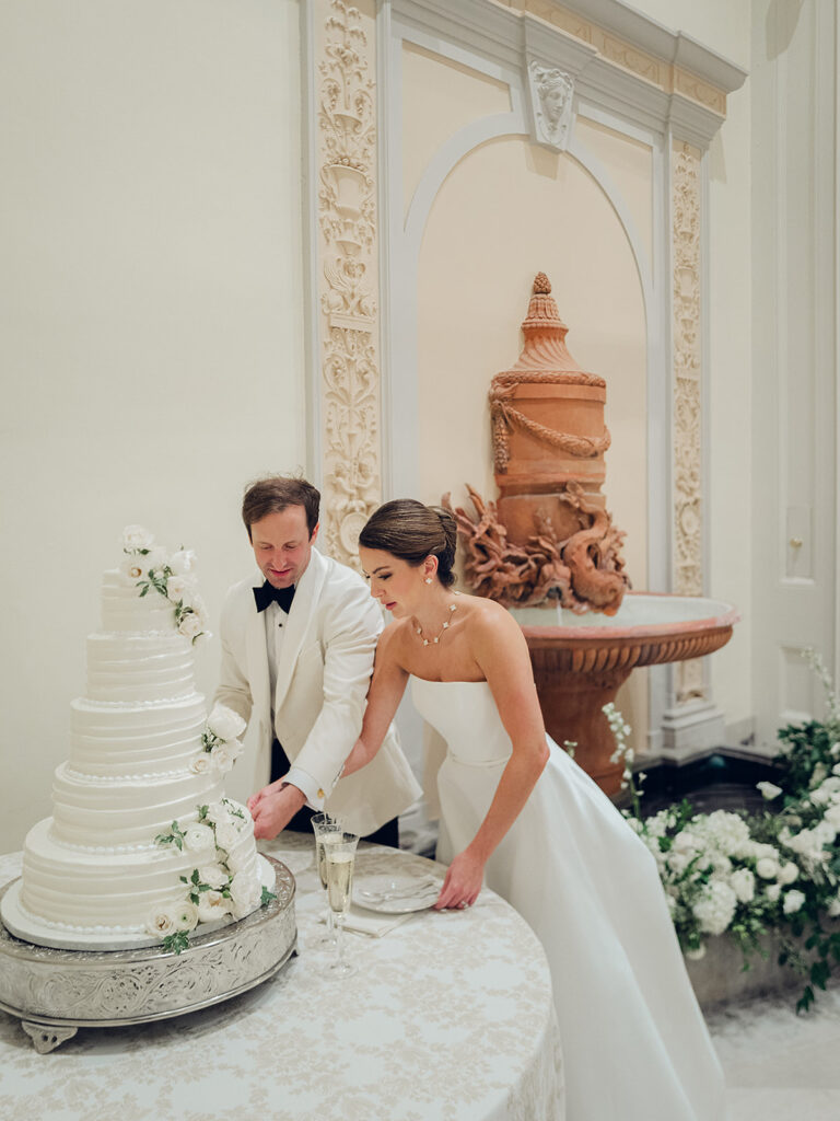 Simple, all-white florals cascade down the side of stunning reception wedding cake. Spray roses, cloud roses, ranunculus, riceflower, olive branches and ivy are used to create a beautiful cake display. Design by Rosemary & Finch Floral Design in Nashville, TN.