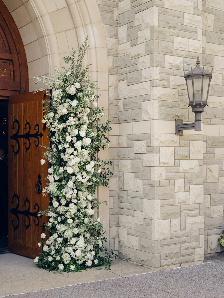Stunning all-white floral arch for wedding ceremony at Covenant Presbyterian Church in Nashville. The arch creates an elegant focal point with flowers consisting of hydrangeas, cloud roses, lisianthus, ranunculus, tulips, dahlias, riceflower, stock, allium, delphinium, and ivy. Meadows with the same flowers surround the bottom of the arch on either side. Design by Rosemary & Finch Floral Design in Nashville, TN.