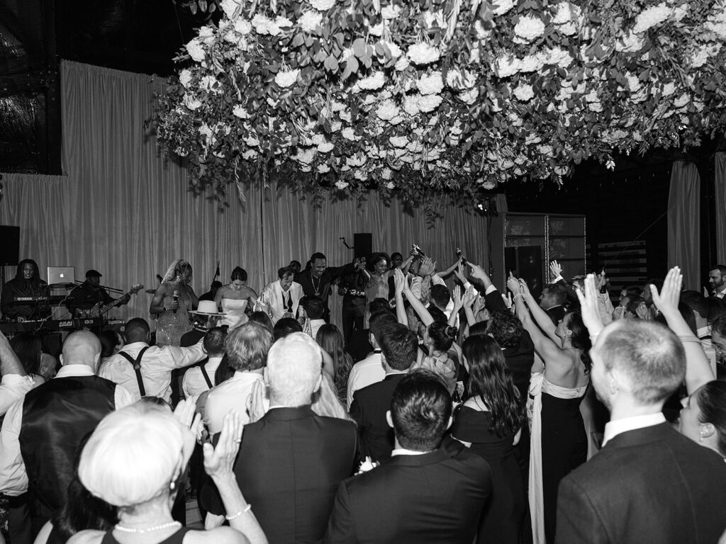 Glamorous, all-white hydrangea floral installation creates a stunning focal point for a reception dance floor. Hundreds of hydrangeas are accompanied with olive branches and other greenery for a lush, romantic feel inside a glass-tented reception at Cheekwood. Garden-like meadows line the stage front consisting of all-white garden roses, hydrangeas, spray roses, riceflower, stock and delphinium. Design by Rosemary & Finch Floral Design in Nashville, TN.