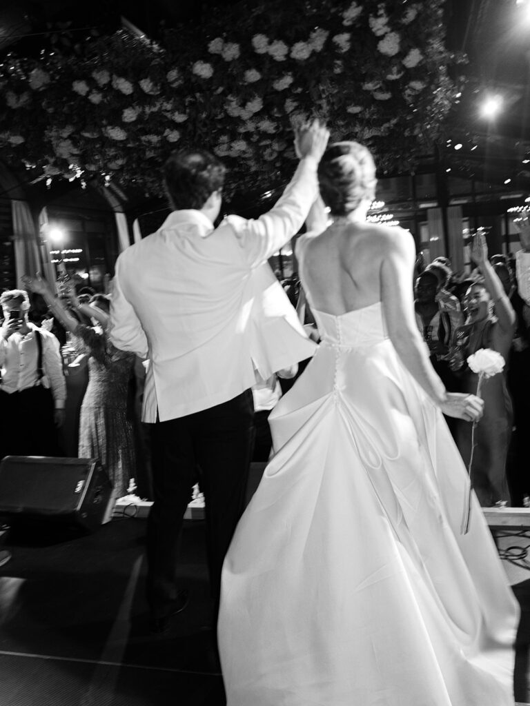 Glamorous, all-white hydrangea floral installation creates a stunning focal point for a reception dance floor. Hundreds of hydrangeas are accompanied with olive branches and other greenery for a lush, romantic feel inside a glass-tented reception at Cheekwood. Garden-like meadows line the stage front consisting of all-white garden roses, hydrangeas, spray roses, riceflower, stock and delphinium. Design by Rosemary & Finch Floral Design in Nashville, TN.