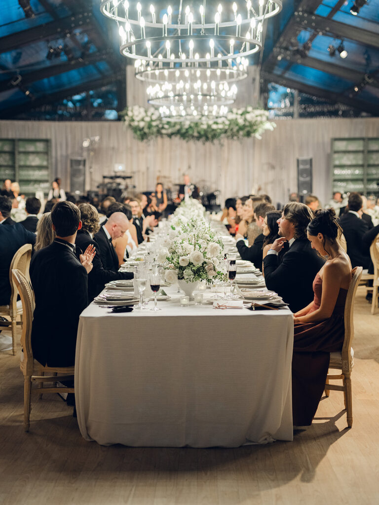 Romantic and stunning glass-tented wedding reception at Cheekwood botanical garden in Nashville, TN. Elegant garden-inspired centerpieces feature lush, all-white cloud roses, hydrangeas, lisianthus, dahlias, sweet peas, double tulips, delphinium, stock, allium, flowering champagne, and more with light greenery. Matching banquette meadows line either side of the glass tent creating a lush and timeless feel. Design by Rosemary & Finch Floral Design in Nashville, TN.