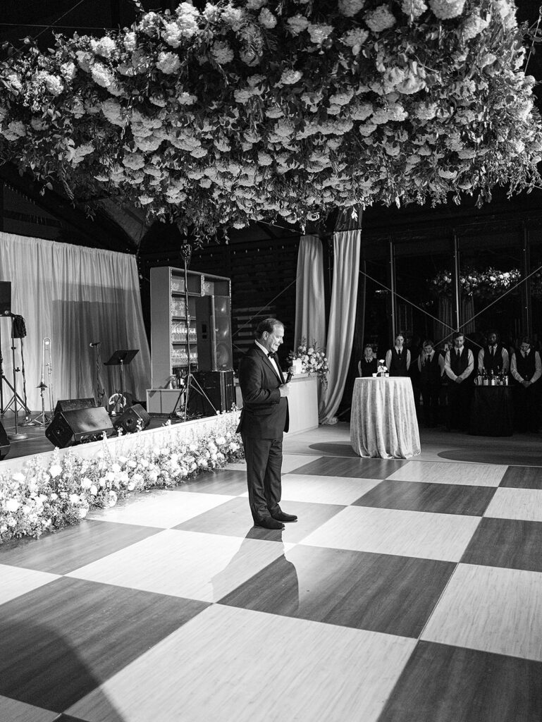 Glamorous, all-white hydrangea floral installation creates a stunning focal point for a reception dance floor. Hundreds of hydrangeas are accompanied with olive branches and other greenery for a lush, romantic feel inside a glass-tented reception at Cheekwood. Garden-like meadows line the stage front consisting of all-white garden roses, hydrangeas, spray roses, riceflower, stock and delphinium. Design by Rosemary & Finch Floral Design in Nashville, TN.