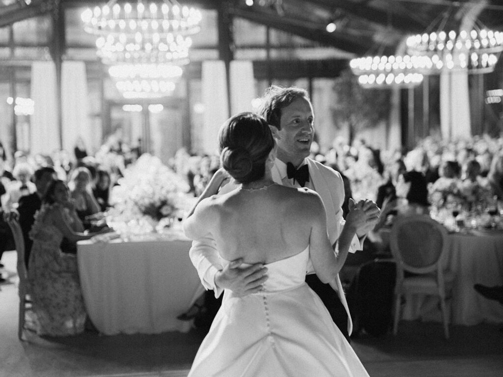 Romantic and stunning glass-tented wedding reception at Cheekwood botanical garden in Nashville, TN. Elegant garden-inspired centerpieces feature lush, all-white cloud roses, hydrangeas, lisianthus, dahlias, sweet peas, double tulips, delphinium, stock, allium, flowering champagne, and more with light greenery. Matching banquette meadows line either side of the glass tent creating a lush and timeless feel. Design by Rosemary & Finch Floral Design in Nashville, TN.