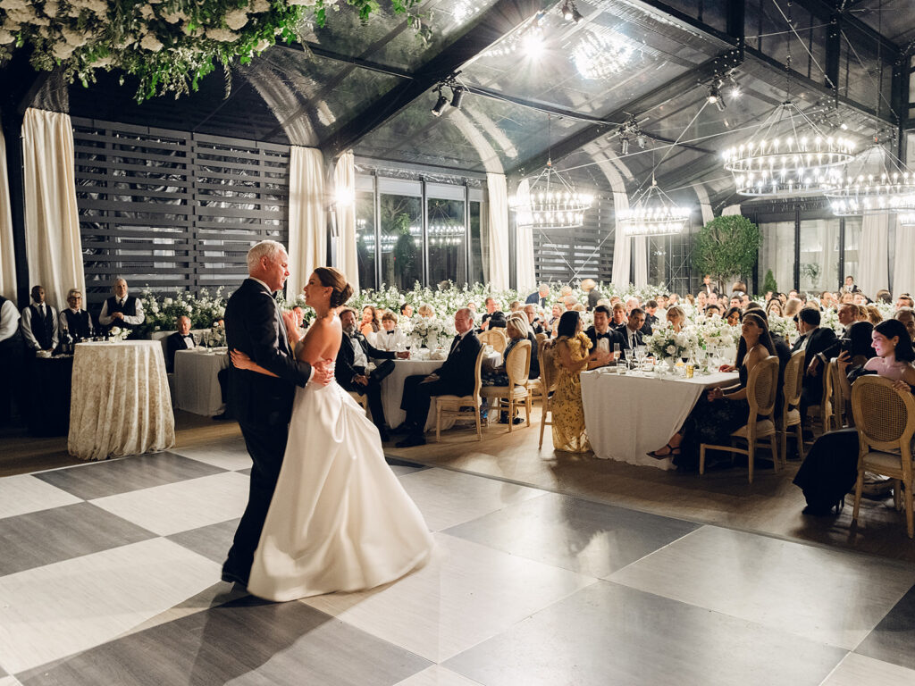 Romantic and stunning glass-tented wedding reception at Cheekwood botanical garden in Nashville, TN. Elegant garden-inspired centerpieces feature lush, all-white cloud roses, hydrangeas, lisianthus, dahlias, sweet peas, double tulips, delphinium, stock, allium, flowering champagne, and more with light greenery. Matching banquette meadows line either side of the glass tent creating a lush and timeless feel. Design by Rosemary & Finch Floral Design in Nashville, TN.