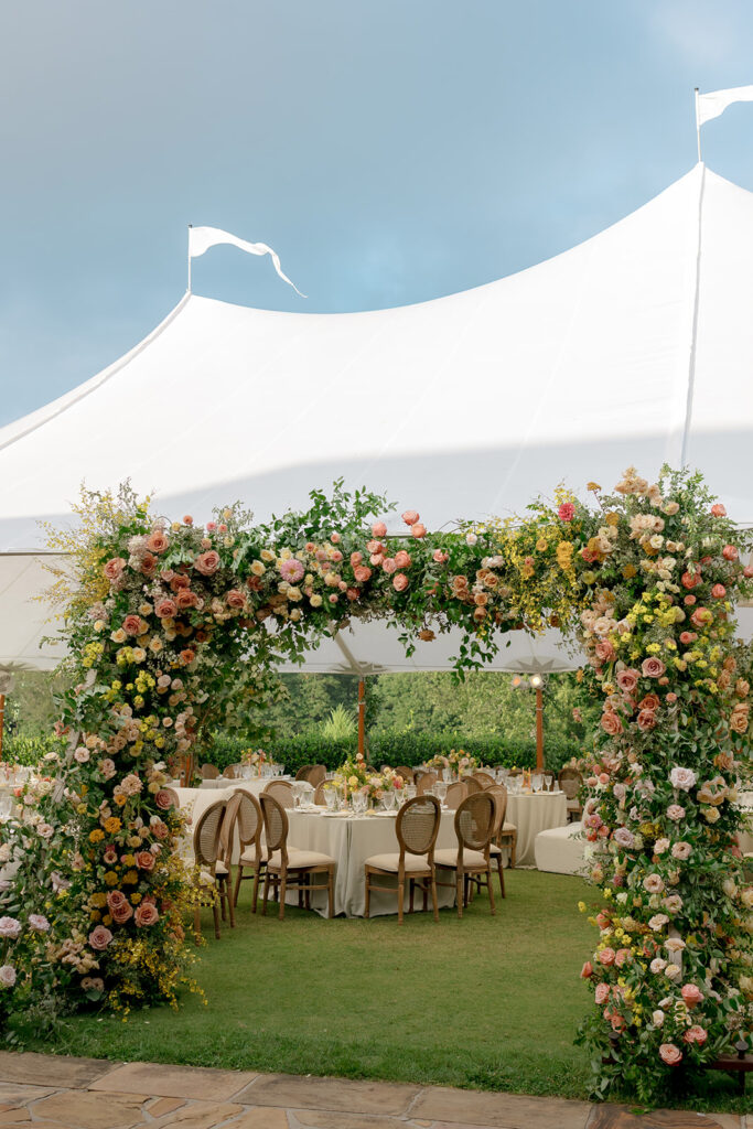 A lush, colorful floral arch frames an elegant tented wedding in Sewanee, TN; Overflowing with garden roses, spray roses, ranunculus, sweet peas, moab roses, lisianthus, yarrow, zinnias, tulips, butterfly ranunculus, foxglove, smokebush, oncidium and greenery in soft shades of blush, peach, butter-yellow and apricot. The floral arch curates a romantic, garden-inspired atmosphere with natural, airy textures and timeless charm. Design by Rosemary and Finch Floral Design in Nashville, TN.