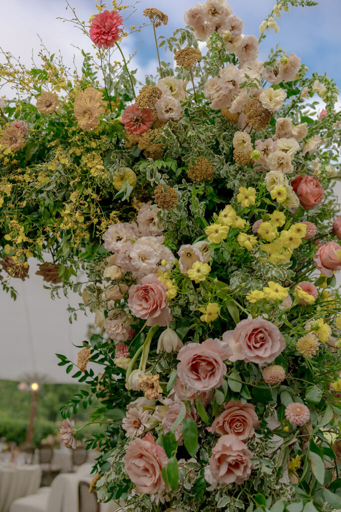 A lush, colorful floral arch frames an elegant tented wedding in Sewanee, TN; Overflowing with garden roses, spray roses, ranunculus, sweet peas, moab roses, lisianthus, yarrow, zinnias, tulips, butterfly ranunculus, foxglove, smokebush, oncidium and greenery in soft shades of blush, peach, butter-yellow and apricot. The floral arch curates a romantic, garden-inspired atmosphere with natural, airy textures and timeless charm. Design by Rosemary and Finch Floral Design in Nashville, TN.