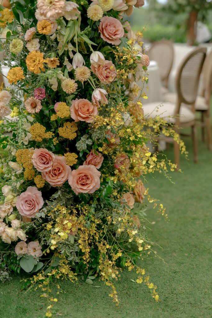 A lush, colorful floral arch frames an elegant tented wedding in Sewanee, TN; Overflowing with garden roses, spray roses, ranunculus, sweet peas, moab roses, lisianthus, yarrow, zinnias, tulips, butterfly ranunculus, foxglove, smokebush, oncidium and greenery in soft shades of blush, peach, butter-yellow and apricot. The floral arch curates a romantic, garden-inspired atmosphere with natural, airy textures and timeless charm. Design by Rosemary and Finch Floral Design in Nashville, TN.