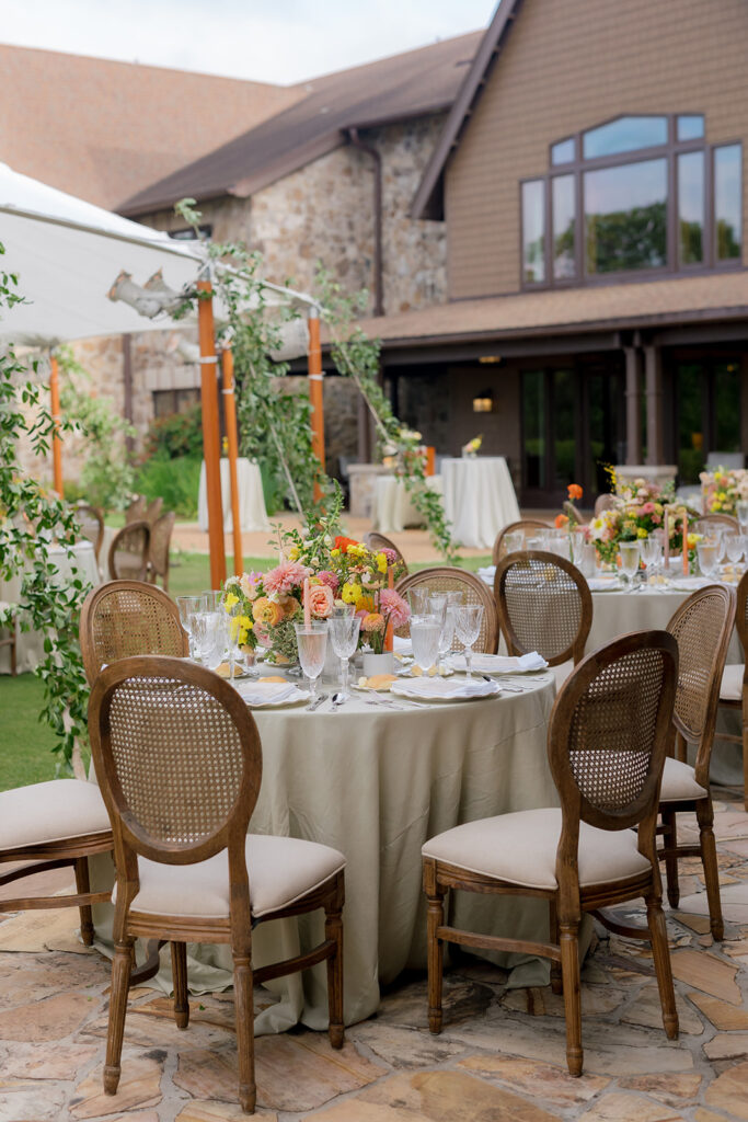 A mix of lush, garden-like floral centerpieces create a colorful display across tables under a canvas tent for a Sewanee wedding reception. Compotes, bud vases and ikebana arrangements overflow with garden roses, moab roses, icelandic poppies, lisianthus, tulips, dahlias, ranunculus, scabiosa, zinnias, sweet peas, oncidium and more in shades of peach, red, apricot and yellow. Lush greenery is suspended around light pendants under the tent. Design by Rosemary and Finch Floral Design in Nashville, TN.
