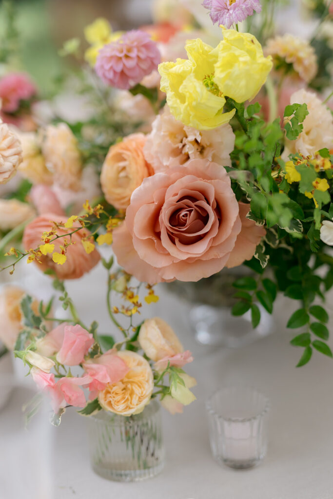 A mix of lush, garden-like floral centerpieces create a colorful display across tables under a canvas tent for a Sewanee wedding. Compotes, bud vases and ikebana arrangements overflow with garden roses, moab roses, icelandic poppies, lisianthus, tulips, dahlias, ranunculus, scabiosa, zinnias, sweet peas, oncidium and more in shades of peach, red, apricot and yellow. Lush greenery is suspended around light pendants under the tent. Design by Rosemary and Finch Floral Design in Nashville, TN.
