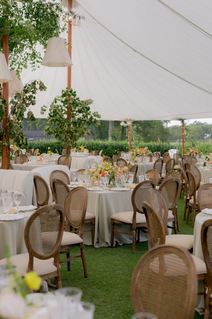 A mix of lush, garden-like floral centerpieces create a colorful display across tables under a canvas tent for a Sewanee wedding reception. Compotes, bud vases and ikebana arrangements overflow with garden roses, moab roses, icelandic poppies, lisianthus, tulips, dahlias, ranunculus, scabiosa, zinnias, sweet peas, oncidium and more in shades of peach, red, apricot and yellow. Lush greenery is suspended around light pendants under the tent. Design by Rosemary and Finch Floral Design in Nashville, TN.
