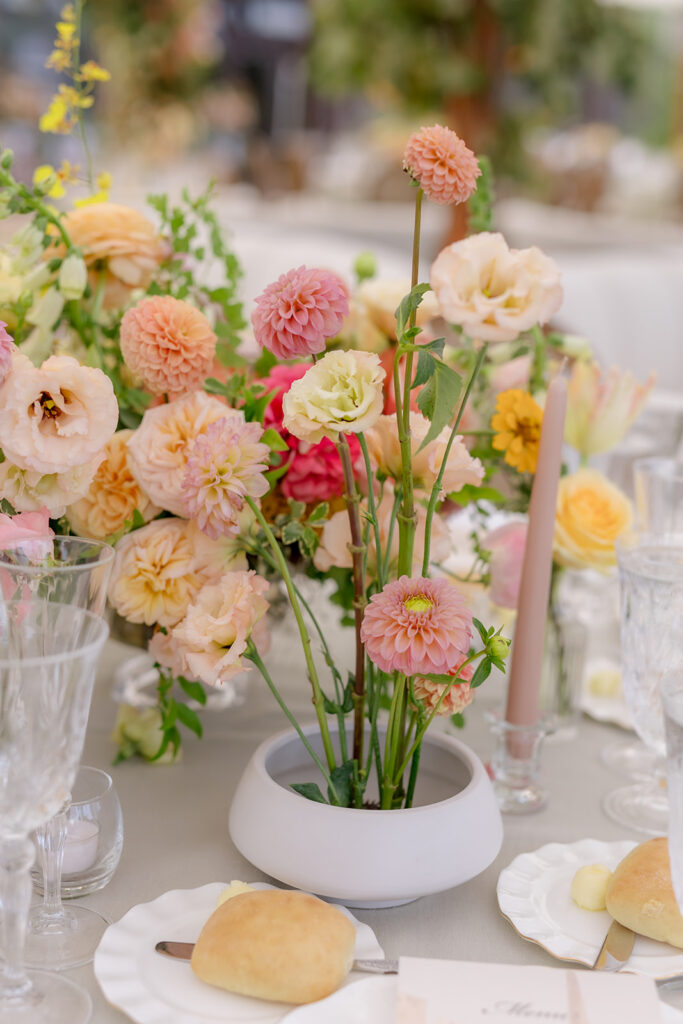 A mix of lush, garden-like floral centerpieces create a colorful display across tables under a canvas tent for a Sewanee wedding reception. Compotes, bud vases and ikebana arrangements overflow with garden roses, moab roses, icelandic poppies, lisianthus, tulips, dahlias, ranunculus, scabiosa, zinnias, sweet peas, oncidium and more in shades of peach, red, apricot and yellow. Lush greenery is suspended around light pendants under the tent. Design by Rosemary and Finch Floral Design in Nashville, TN.
