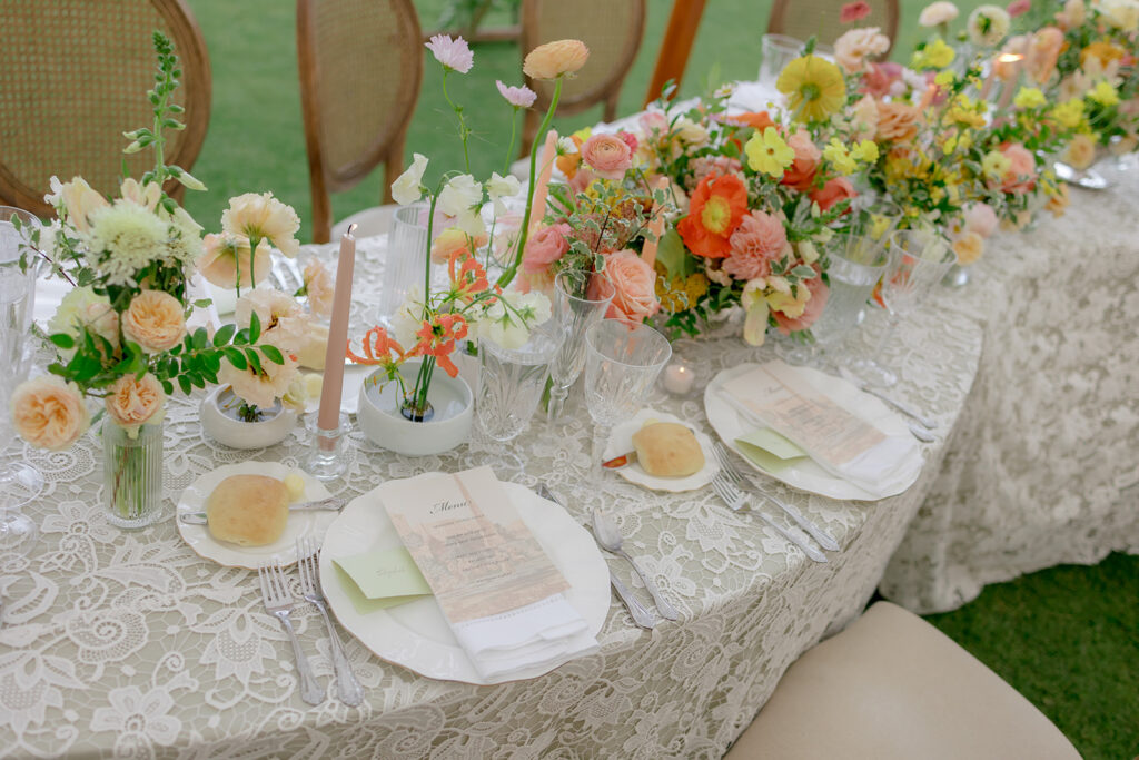 A mix of lush, garden-like floral centerpieces create a colorful display across tables under a canvas tent for a Sewanee wedding. Compotes, bud vases and ikebana arrangements overflow with garden roses, moab roses, icelandic poppies, lisianthus, tulips, dahlias, ranunculus, scabiosa, zinnias, sweet peas, oncidium and more in shades of peach, red, apricot and yellow. Lush greenery is suspended around light pendants under the tent. Design by Rosemary and Finch Floral Design in Nashville, TN.
