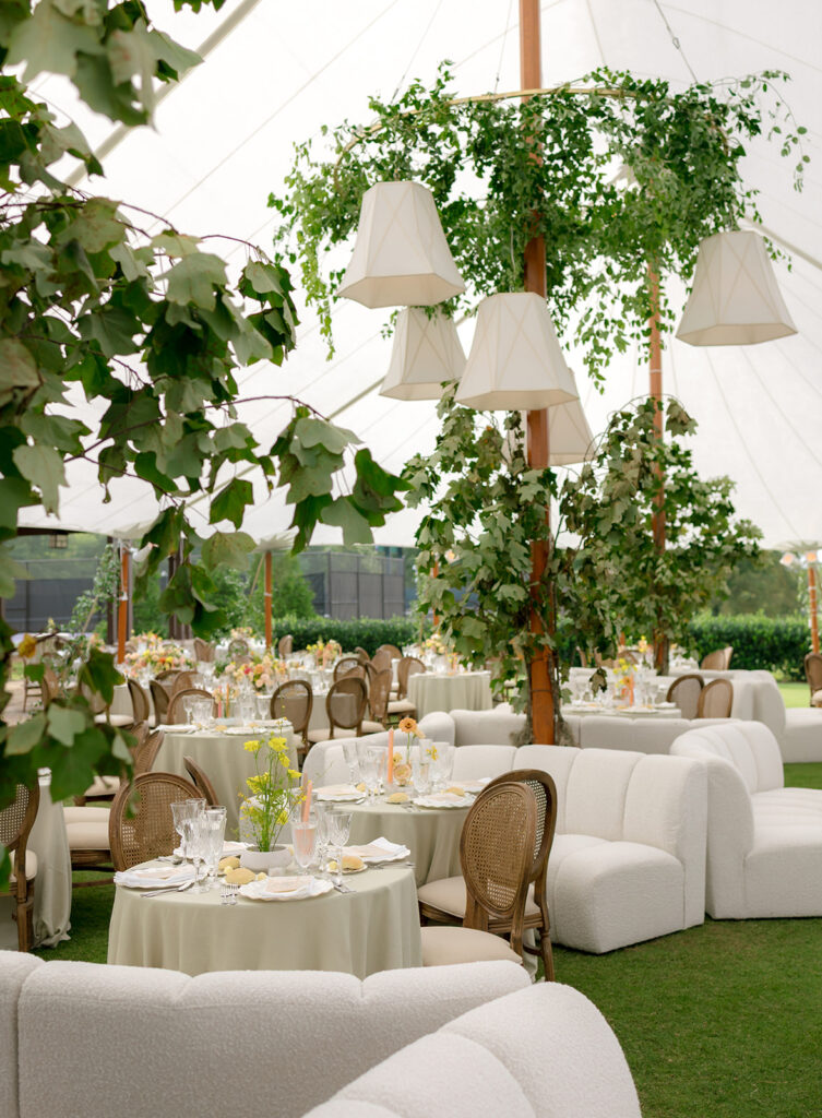 A mix of lush, garden-like floral centerpieces create a colorful display across tables under a canvas tent for a Sewanee wedding. Compotes, bud vases and ikebana arrangements overflow with garden roses, moab roses, icelandic poppies, zinnias, sweet peas, oncidium and more in shades of peach, red, apricot and yellow. Lush greenery is suspended around light pendants and wrapped around tent poles under the tent. Design by Rosemary and Finch Floral Design in Nashville, TN.
