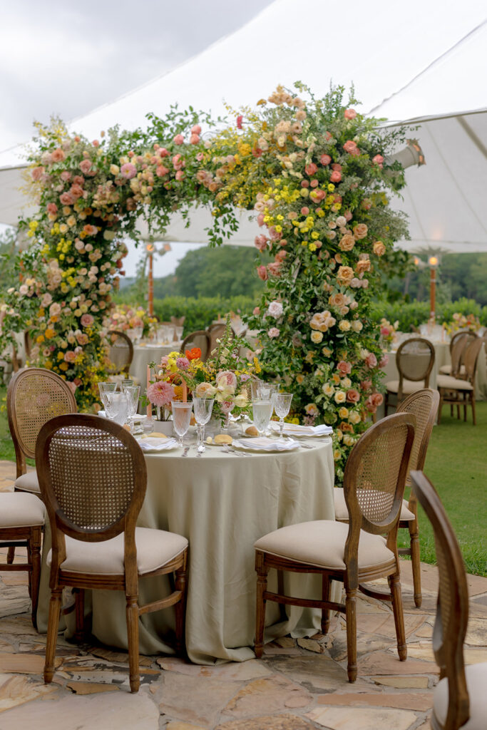 A lush, colorful floral arch frames an elegant tented wedding in Sewanee, TN; Overflowing with garden roses, spray roses, ranunculus, sweet peas, moab roses, lisianthus, yarrow, zinnias, tulips, butterfly ranunculus, foxglove, smokebush, oncidium and greenery in soft shades of blush, peach, butter-yellow and apricot. The floral arch curates a romantic, garden-inspired atmosphere with natural, airy textures and timeless charm. Design by Rosemary and Finch Floral Design in Nashville, TN.
