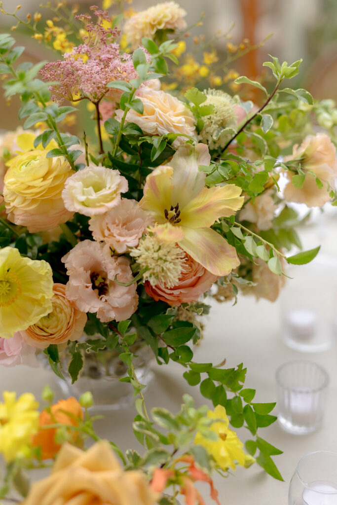 A mix of lush, garden-like floral centerpieces create a colorful display across tables under a canvas tent for a Sewanee wedding reception. Compotes, bud vases and ikebana arrangements overflow with garden roses, moab roses, icelandic poppies, lisianthus, tulips, dahlias, ranunculus, scabiosa, zinnias, sweet peas, oncidium and more in shades of peach, red, apricot and yellow. Lush greenery is suspended around light pendants under the tent. Design by Rosemary and Finch Floral Design in Nashville, TN.
