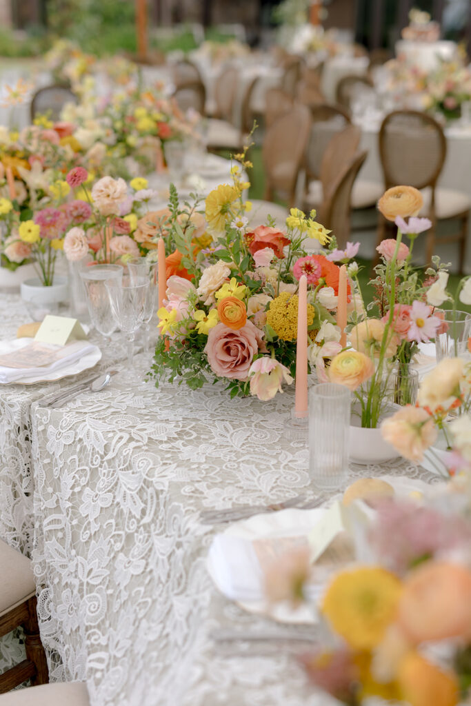 A mix of lush, garden-like floral centerpieces create a colorful display across tables under a canvas tent for a Sewanee wedding reception. Compotes, bud vases and ikebana arrangements overflow with garden roses, moab roses, icelandic poppies, lisianthus, tulips, dahlias, ranunculus, scabiosa, zinnias, sweet peas, oncidium and more in shades of peach, red, apricot and yellow. Lush greenery is suspended around light pendants under the tent. Design by Rosemary and Finch Floral Design in Nashville, TN.
