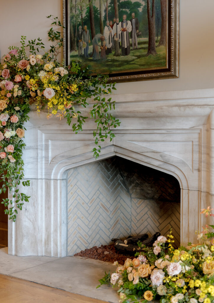 Stunning floral installation with bright colors of yellow, peach, blush, and green, perfectly frame fireplace in the Sewanee Inn. Flowers highlighted are garden roses, spray roses, moab roses, butterfly ranunculus, ranunculus, lisianthus, zinnias, foxglove, gloriosa, oncidium, smokebush,  cottage yarrow, carnations, tulips and greenery including smilax. The wedding cake florals are also highlighted near the fireplace. Design by Rosemary and Finch Floral Design in Nashville, TN.
