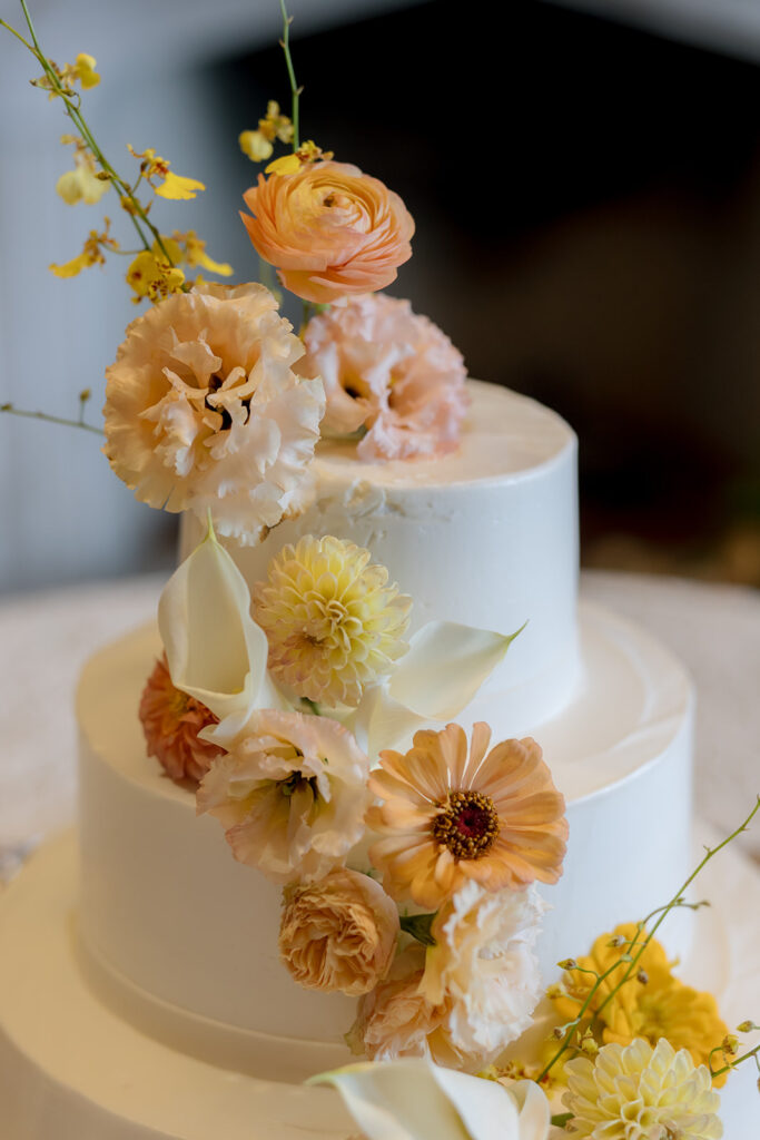 Stunning floral installation with bright colors of yellow, peach, blush, and green, perfectly frame fireplace in the Sewanee Inn. Flowers highlighted are garden roses, spray roses, moab roses, butterfly ranunculus, ranunculus, lisianthus, zinnias, foxglove, gloriosa, oncidium, smokebush,  cottage yarrow, carnations, tulips and greenery including smilax. The wedding cake florals are also highlighted near the fireplace. Design by Rosemary and Finch Floral Design in Nashville, TN.