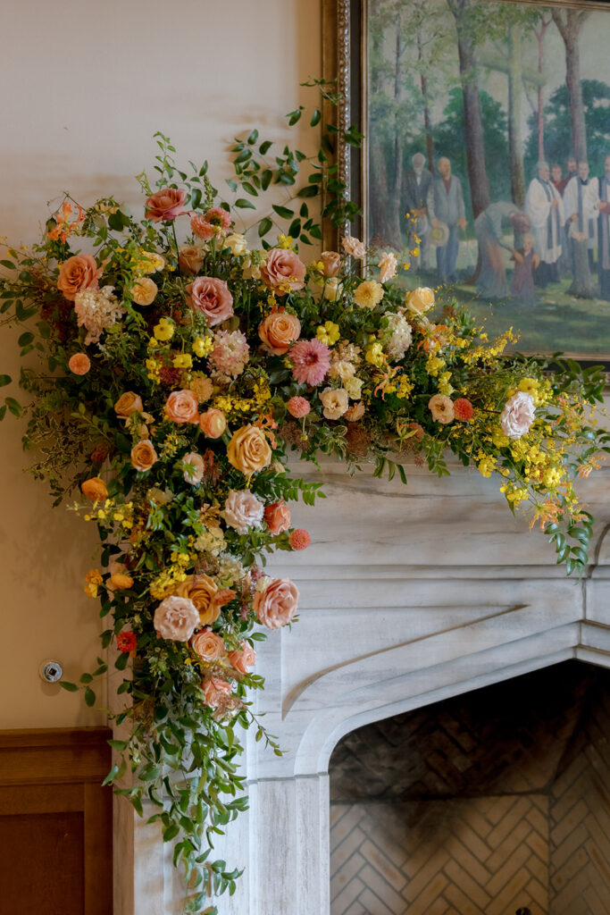 Stunning floral installation with bright colors of yellow, peach, blush, and green, perfectly frame fireplace in the Sewanee Inn. Flowers highlighted are garden roses, spray roses, moab roses, butterfly ranunculus, ranunculus, lisianthus, zinnias, foxglove, gloriosa, oncidium, smokebush,  cottage yarrow, carnations, tulips and greenery including smilax. The wedding cake florals are also highlighted near the fireplace. Design by Rosemary and Finch Floral Design in Nashville, TN.