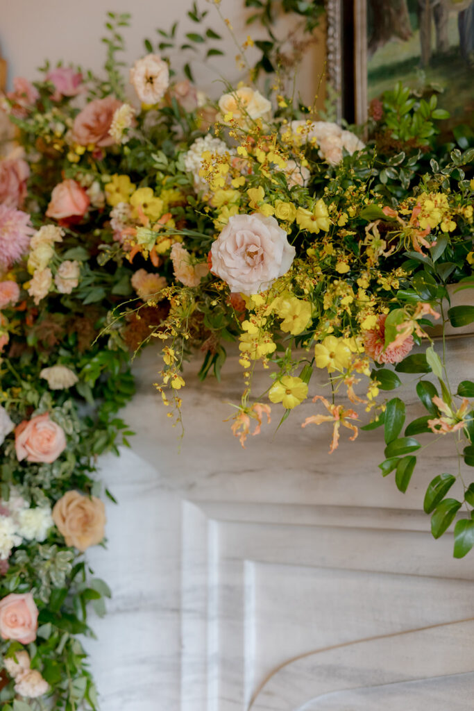 Stunning floral installation with bright colors of yellow, peach, blush, and green, perfectly frame fireplace in the Sewanee Inn. Flowers highlighted are garden roses, spray roses, moab roses, butterfly ranunculus, ranunculus, lisianthus, zinnias, foxglove, gloriosa, oncidium, smokebush,  cottage yarrow, carnations, tulips and greenery including smilax. The wedding cake florals are also highlighted near the fireplace. Design by Rosemary and Finch Floral Design in Nashville, TN.