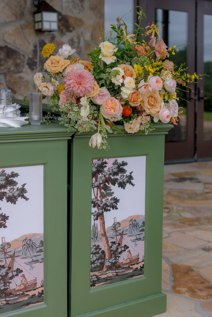 Stunning reception and bar statement arrangements catch the eye of guests at Sewanee, TN wedding. Colorful bursts of garden roses, moab roses, creme de la creme, carnations, spray roses, dahlias, tulips, yarrow, foxglove, zinnias, oncidium orchids, cosmos, smokebush and more are accompanied by soft greens and ferns. Arrangements are drenched in colors of peach, pinks, yellows and green. Design by Rosemary and Finch Floral Design in Nashville, TN. 