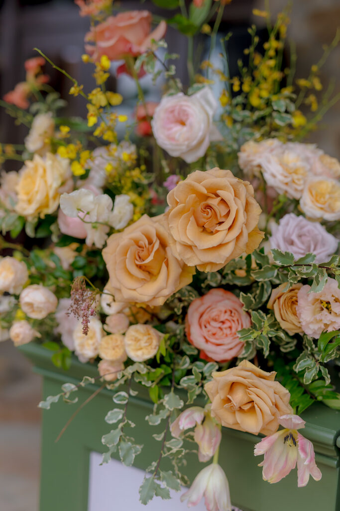 Stunning reception and bar statement arrangements catch the eye of guests at Sewanee, TN wedding. Colorful bursts of garden roses, moab roses, creme de la creme, carnations, spray roses, dahlias, tulips, yarrow, foxglove, zinnias, oncidium orchids, cosmos, smokebush and more are accompanied by soft greens and ferns. Arrangements are drenched in colors of peach, pinks, yellows and green. Design by Rosemary and Finch Floral Design in Nashville, TN. 