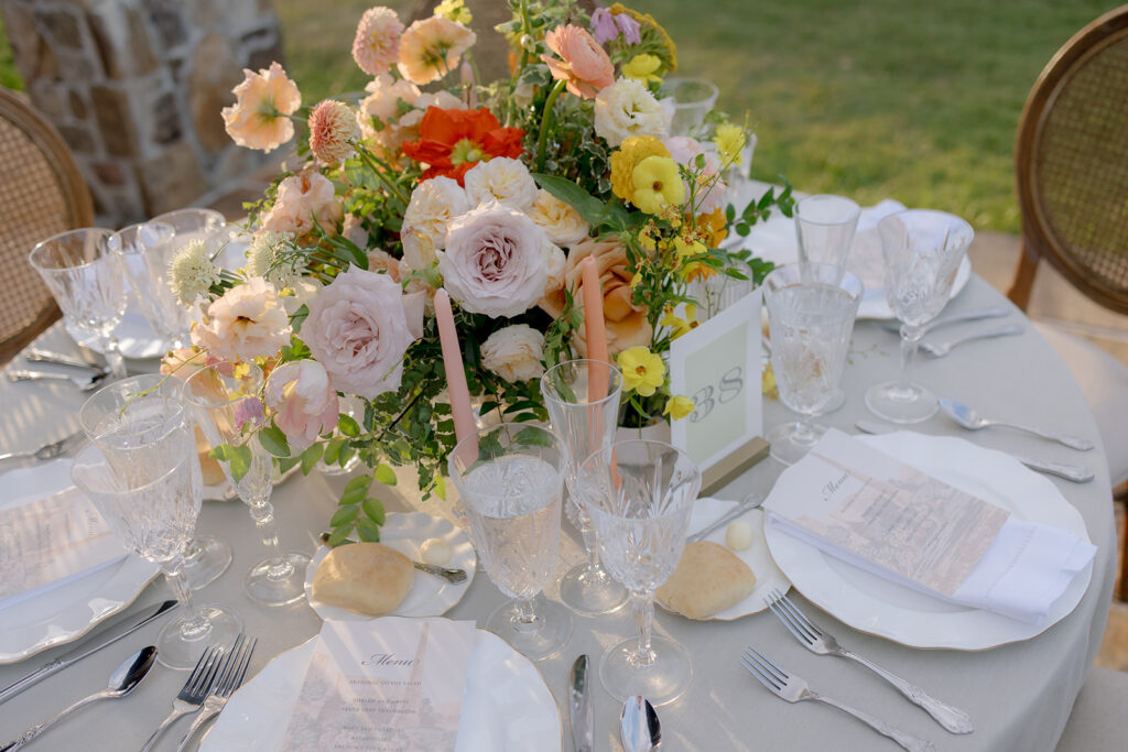 A mix of lush, garden-like floral centerpieces create a colorful display across tables under a canvas tent for a Sewanee wedding reception. Compotes, bud vases and ikebana arrangements overflow with garden roses, moab roses, icelandic poppies, lisianthus, tulips, dahlias, ranunculus, scabiosa, zinnias, sweet peas, oncidium and more in shades of peach, red, apricot and yellow. Lush greenery is suspended around light pendants under the tent. Design by Rosemary and Finch Floral Design in Nashville, TN.
