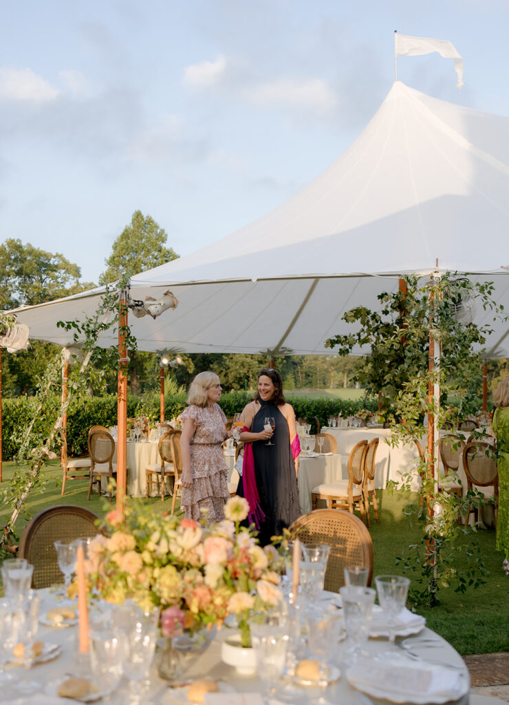 A mix of lush, garden-like floral centerpieces create a colorful display across tables under a canvas tent for a Sewanee wedding reception. Compotes, bud vases and ikebana arrangements overflow with garden roses, moab roses, icelandic poppies, lisianthus, tulips, dahlias, ranunculus, scabiosa, zinnias, sweet peas, oncidium and more in shades of peach, red, apricot and yellow. Lush greenery is suspended around light pendants under the tent. Design by Rosemary and Finch Floral Design in Nashville, TN.
