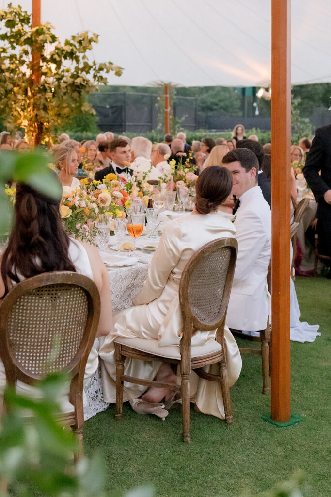 A mix of lush, garden-like floral centerpieces create a colorful display across tables under a canvas tent for a Sewanee wedding reception. Compotes, bud vases and ikebana arrangements overflow with garden roses, moab roses, icelandic poppies, lisianthus, tulips, dahlias, ranunculus, scabiosa, zinnias, sweet peas, oncidium and more in shades of peach, red, apricot and yellow. Lush greenery is suspended around light pendants under the tent. Design by Rosemary and Finch Floral Design in Nashville, TN.
