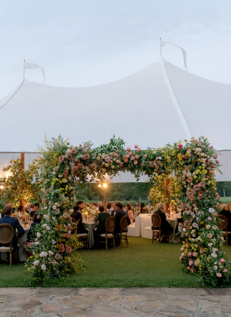 A lush, colorful floral arch frames an elegant tented wedding in Sewanee, TN; Overflowing with garden roses, spray roses, ranunculus, sweet peas, moab roses, lisianthus, yarrow, zinnias, tulips, butterfly ranunculus, foxglove, smokebush, oncidium and greenery in soft shades of blush, peach, butter-yellow and apricot. The floral arch curates a romantic, garden-inspired atmosphere with natural, airy textures and timeless charm. Design by Rosemary and Finch Floral Design in Nashville, TN.