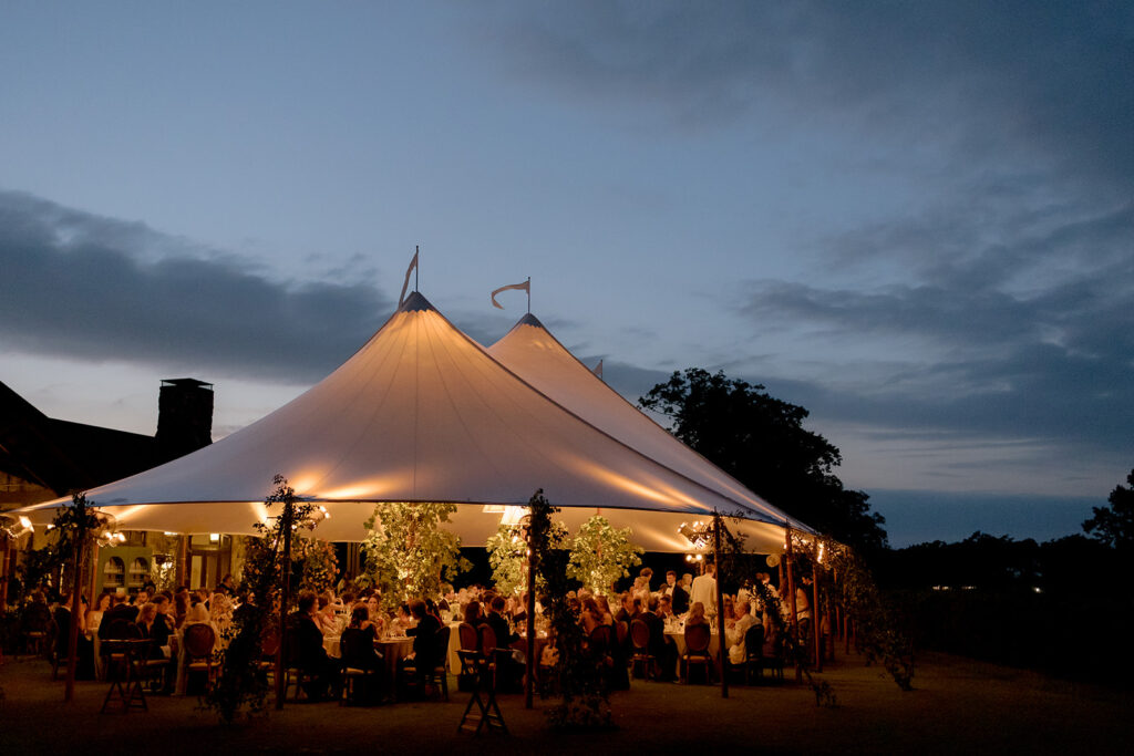 A mix of lush, garden-like floral centerpieces create a colorful display across tables under a canvas tent for a Sewanee wedding reception. Compotes, bud vases and ikebana arrangements overflow with garden roses, moab roses, icelandic poppies, lisianthus, tulips, dahlias, ranunculus, scabiosa, zinnias, sweet peas, oncidium and more in shades of peach, red, apricot and yellow. Lush greenery is suspended around light pendants under the tent. Design by Rosemary and Finch Floral Design in Nashville, TN.
